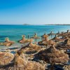 Spiaggia con ombrelloni di paglia, mare calmo e isola in lontananza.
