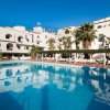 Edificio bianco a tre piani con piscina riflettente, palme e cielo azzurro.