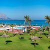 Prato verde con ombrelloni di paglia, palme e piscina che si affaccia sul mare blu.