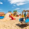Area giochi sulla sabbia con scivoli colorati e strutture per bambini sotto un cielo azzurro.